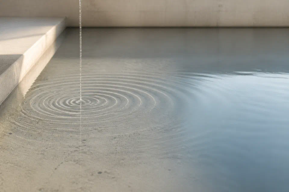 A continuous narrow stream falls onto calm water, creating evenly spreading concentric ripples